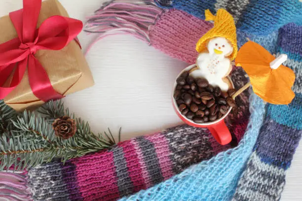 cheerful gingerbread snowman sits under an umbrella in a cup with coffee beans, next to a gift, knitted scarves and a spruce branch top view