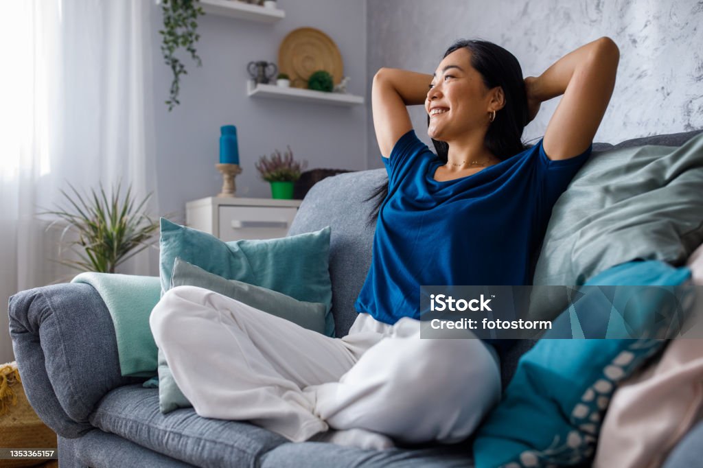 Copy space shot of young woman lounging on sofa with hands behind head and daydreaming Full length shot of beautiful, happy young woman sitting on the cozy sofa with hands behind head, looking away, smiling and daydreaming. Relaxation Stock Photo