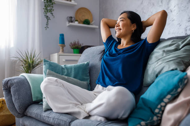 Copy space shot of young woman lounging on sofa with hands behind head and daydreaming Full length shot of beautiful, happy young woman sitting on the cozy sofa with hands behind head, looking away, smiling and daydreaming. relaxation stock pictures, royalty-free photos & images