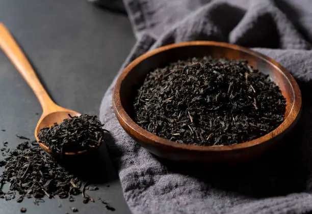 Photo of Tea leaves in a wooden plate and spoon on a black background.