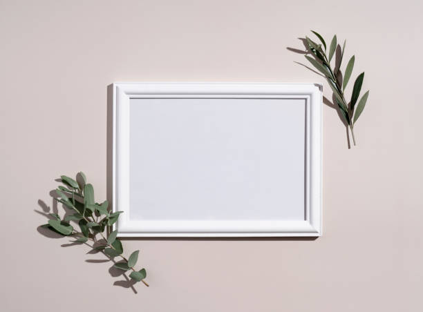 istock Picture frame mockup with white wooden frame. Eucalyptus leaves on beige background. Natural light and shadow overlay. Flat lay, top view. 1353366782