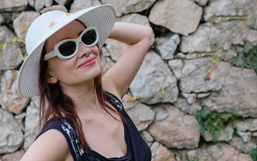 A close up shot of a sweet caucasian lady with hat and sunglasses with her back to a stone wall.
