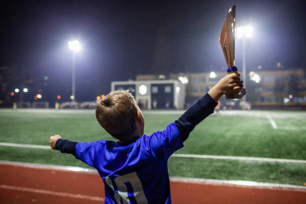 Young soccer player in blue jersey with ten number holding a winners cup after the winning goal Young soccer player in blue jersey with ten number raising a trophy after the winning goal in football competition, illuminated stadium, sport, winner and success. Dreams come true. Rear back view celebrity fans stock pictures, royalty-free photos & images