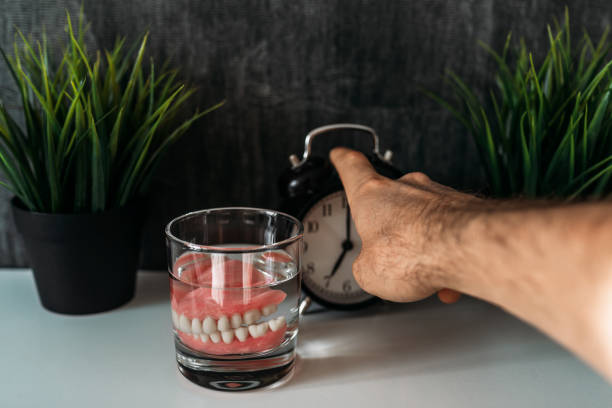 Removable jaw in a glass of cleaning liquid in the early morning. A man's hand, false teeth in a glass with a solution, a watch. Dental prosthesis care. An elderly man turns off the alarm clock stock photo