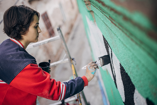 One woman, young female street artist standing on scaffolding and painting on a building wall outdoors.