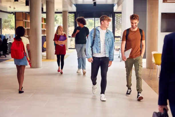 Two Male Students In Busy University Or College Building Talking As They Walk Along Corridor