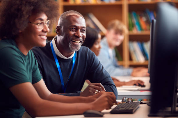 istock Male University Or College Student Working At Computer In Library Being Helped By Tutor 1353372068