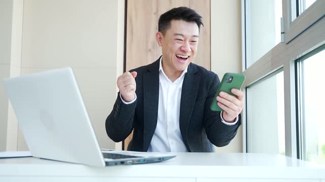 Happy young Asian business man in office looking at mobile phone, with emotion winner or win, financial stock sports betting.