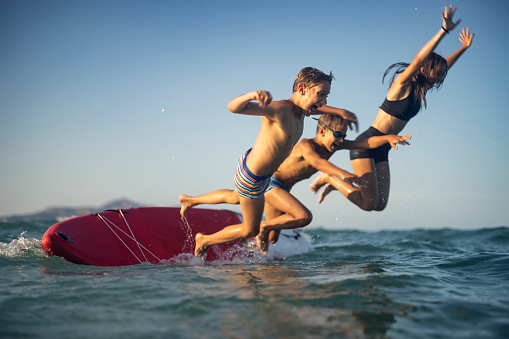 Teenage girl and her brothers are enjoying summer vacations. They are jumping off the the SUP paddleboard into the sea. The setting sun is shining on the kids with warm sunlight.\nCanon R5