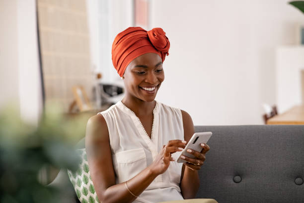 Smiling black woman with african turban using smartphone at home Smiling middle aged african woman with traditional head turban sitting on couch at home using smartphone. Beautiful african american woman with typical headscarf scrolling through internet on cellphone. Happy mid adult lady sitting on sofa browsing mobile phone. turban stock pictures, royalty-free photos & images