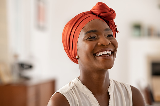 Smiling middle aged African American woman with orange headscarf at home. Beautiful black woman in casual clothing with traditional turban at home laughing. Portrait of mature carefree lady smiling and looking away.