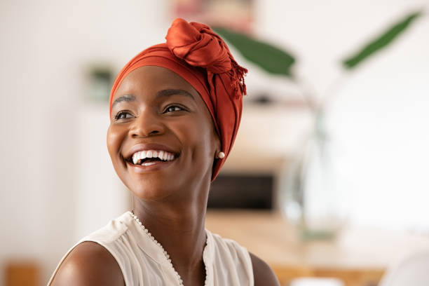 Joyful mature african woman laughing and looking away Beautiful mature black woman with african turban relaxing at home. Cheerful middle aged black woman in casual clothing with traditional headscarf at home laughing. African american lady smiling and looking away with copy space. turban stock pictures, royalty-free photos & images