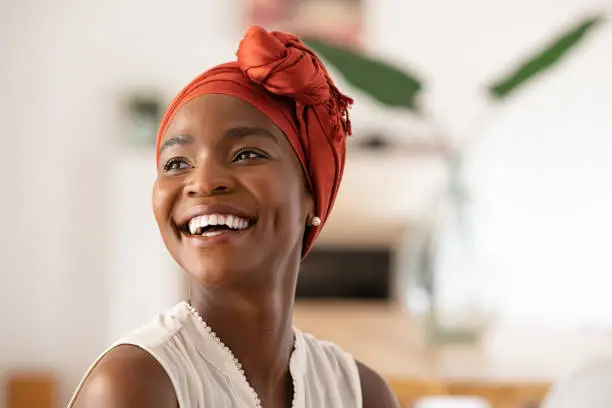 Beautiful mature black woman with african turban relaxing at home. Cheerful middle aged black woman in casual clothing with traditional headscarf at home laughing. African american lady smiling and looking away with copy space.