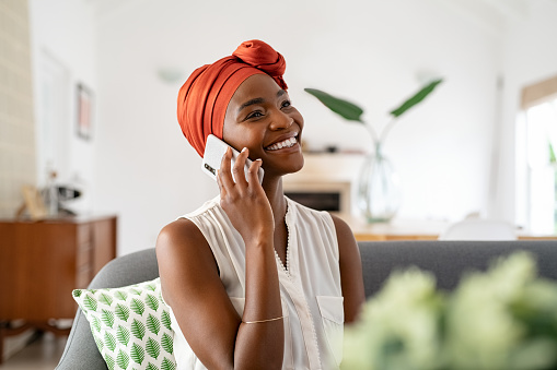 Smiling middle aged woman sitting on couch at home talking over smartphone. Happy black woman with traditional head turban using mobile phone for conversation. Relaxing mid african american lady sitting on sofa at home while using cellphone.