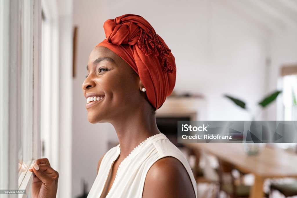 African woman looking out of window while wearing traditional turban Smiling mid adult african american woman with turban looking outside the window. Cheerful black mature woman wearing traditional red headscarf while contempliting outdoor. Happy middle aged lady looking outside the window at home while thinking. Women Stock Photo