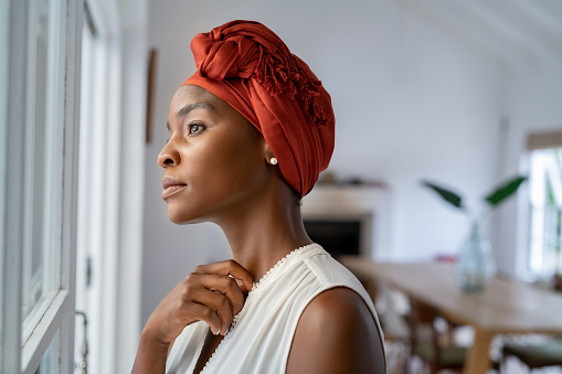 Beautiful pensive woman wearing headscarf looking outside window in contemplation. Mature black woman wearing a traditional turban and standing near window at home while thinking. Worried african mature woman with cancer at home.