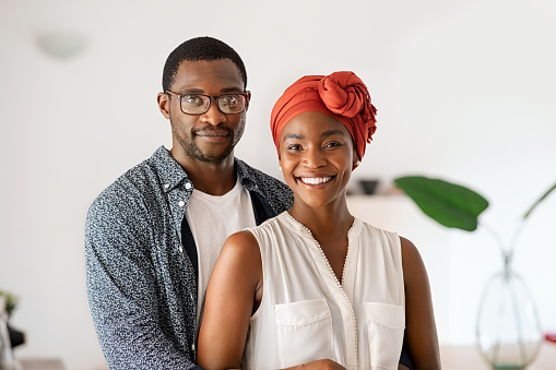 Portrait of cheerful middle aged couple embracing at home. Romantic mid adult black man hugging beautiful wife with traditional red turban while smiling and looking at camera. Happy mature african couple posing.