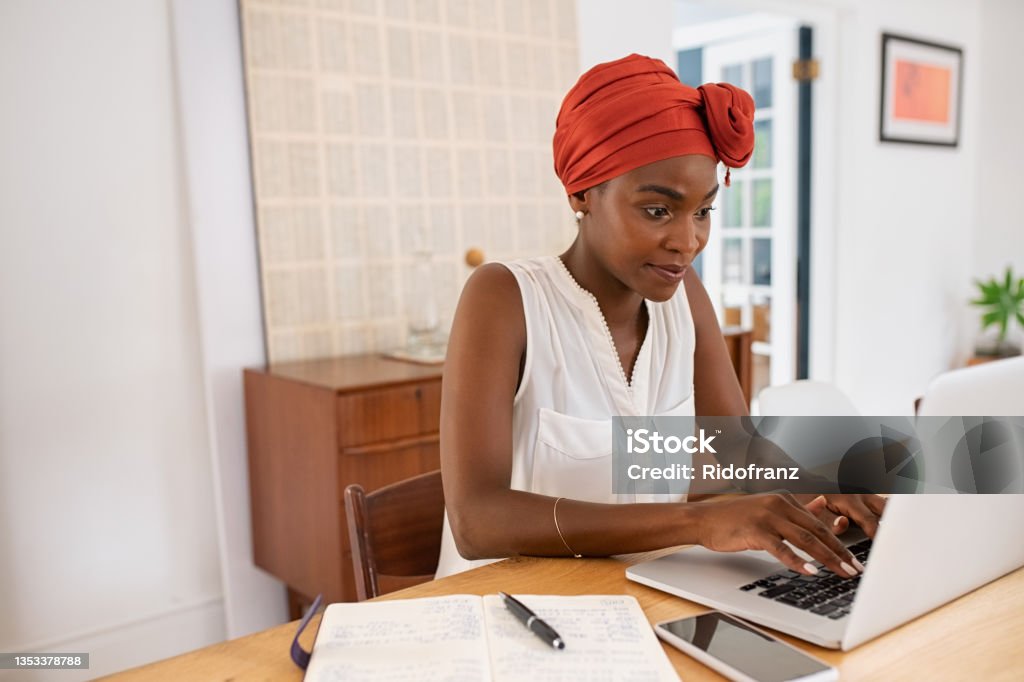 Mature african american woman working on laptop from home Mature black woman with traditional turban sitting at home while working on laptop. Successful black businesswoman typing on laptop wearing a traditional turban while working from home with copy space. Mid adult entrepreneur using computer during lockdown. Women Stock Photo