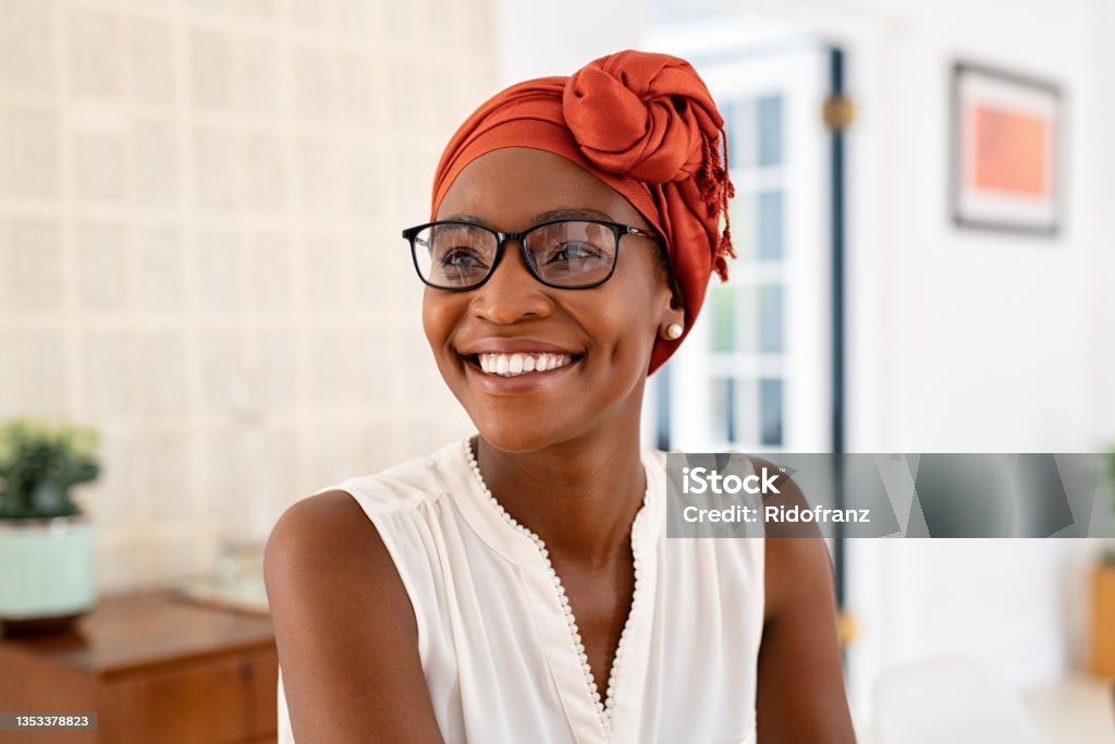 Happy smiling black woman with spectacles wearing african turban Cheerful mature black woman wearing spectacles and traditional turban at home. Beautiful african american woman with headscarf and eyeglasses sitting at home and looking away. Mature lady relaxing at home. Eyeglasses Stock Photo