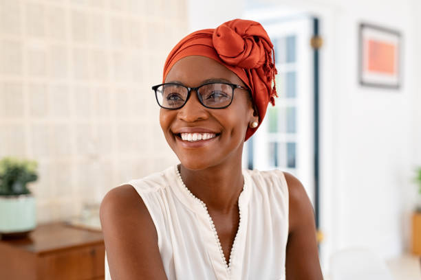 Happy smiling black woman with spectacles wearing african turban Cheerful mature black woman wearing spectacles and traditional turban at home. Beautiful african american woman with headscarf and eyeglasses sitting at home and looking away. Mature lady relaxing at home. beautiful older black woman stock pictures, royalty-free photos & images