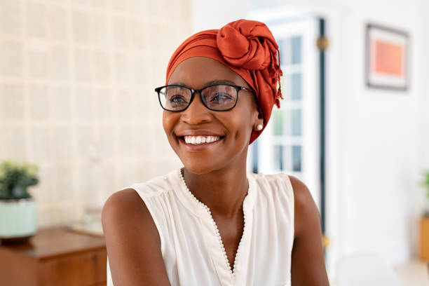 Happy smiling black woman with spectacles wearing african turban Cheerful mature black woman wearing spectacles and traditional turban at home. Beautiful african american woman with headscarf and eyeglasses sitting at home and looking away. Mature lady relaxing at home. typical average stock pictures, royalty-free photos & images