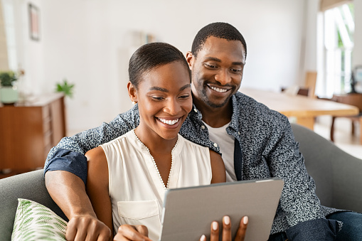 Cheerful black couple sitting on couch and watching movie on digital tablet at home. Mature black husband and beautiful wife using digital tablet to do a video call while sitting on sofa. Middle aged man and african woman relaxing on couch in living room.