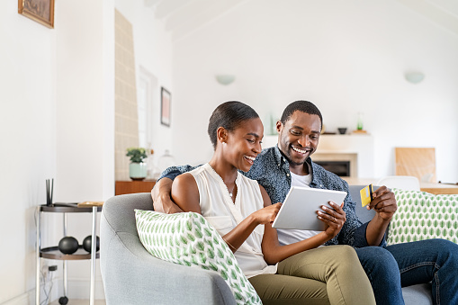 Happy african american wife doing shopping online while using digital tablet and credit card at home with husband. Middle aged black couple relaxing at home while making an online purchase using debit card on digital tablet. Mid couple using bank card to book holidays.