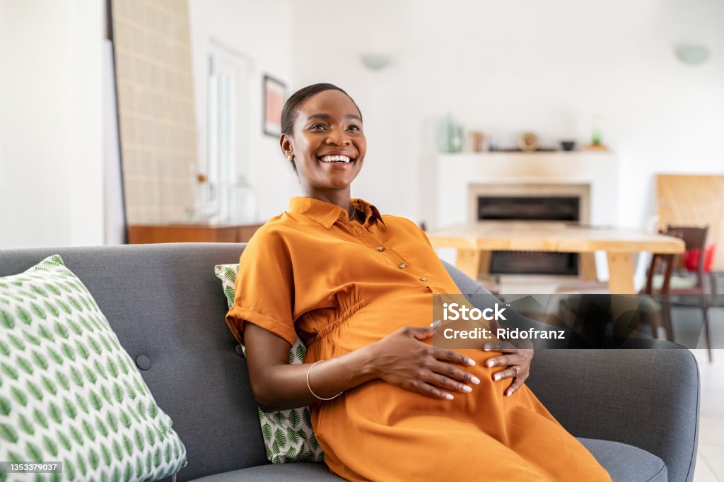Happy mature african pregnant woman smiling at home Portrait of beautiful pregnant woman relaxing at home while touching belly. Smiling middle aged african american woman resting during pregnancy in living room. Happy black pregnant woman sitting in couch and touching her tummy at home. Pregnant Stock Photo