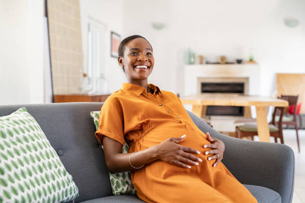 Happy mature african pregnant woman smiling at home Portrait of beautiful pregnant woman relaxing at home while touching belly. Smiling middle aged african american woman resting during pregnancy in living room. Happy black pregnant woman sitting in couch and touching her tummy at home. pregnant stock pictures, royalty-free photos & images