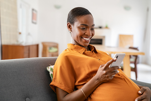 Beautiful mature pregnant woman watching funny video on smartphone. Happy black pregnant woman using mobile while touching tummy at home with copy space. Cheerful middle aged expecting african american woman using cellphone while relaxing on couch at home.