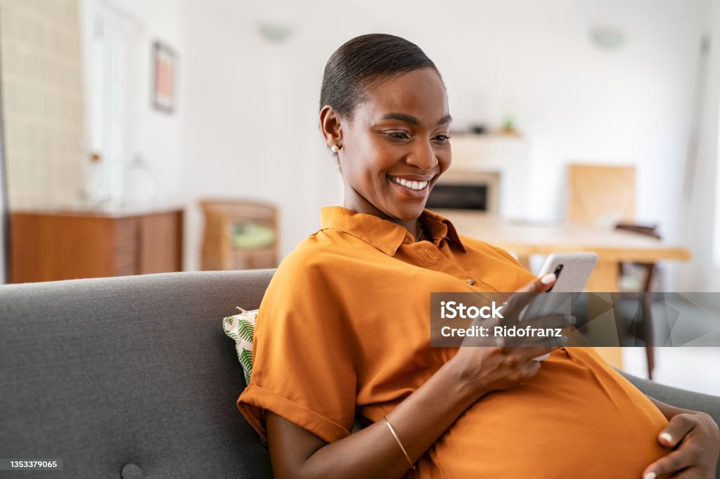 Smiling pregnant mid adult black woman messaging on mobile phone Beautiful mature pregnant woman watching funny video on smartphone. Happy black pregnant woman using mobile while touching tummy at home with copy space. Cheerful middle aged expecting african american woman using cellphone while relaxing on couch at home. Pregnant Stock Photo