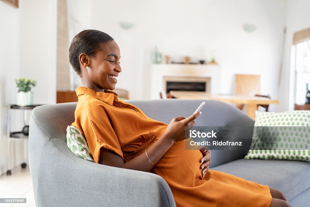 Pregnant african woman using smartphone at home Mature black pregnant woman texting on mobile phone while sitting on couch in living room. Happy mid adult african american pregnant woman using smartphone at home. Beautiful lady messaging on cellphone while relaxing on sofa at home: motherhood and pregnancy concept. Pregnant Stock Photo