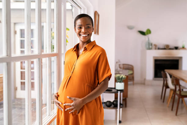 Portrait of happy black pregnant woman looking at camera Happy middle aged pregnant woman with big tummy relaxing at home. Portrait of mature pregnant african american woman standing near the window and caressing her belly. Smiling mid black lady expecting child and looking at camera with copy sapce. surrogacy stock pictures, royalty-free photos & images