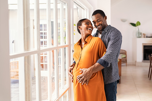 Lovely mid black man hugging his pregnant wife from behind standing near window at home. Happy middle aged black husband embracing pregnant woman while waiting for baby. African american mature couple hugging and looking at each other.
