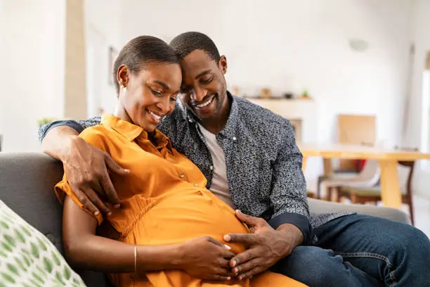 Photo of Black expectant parents sitting on sofa dreaming about their baby