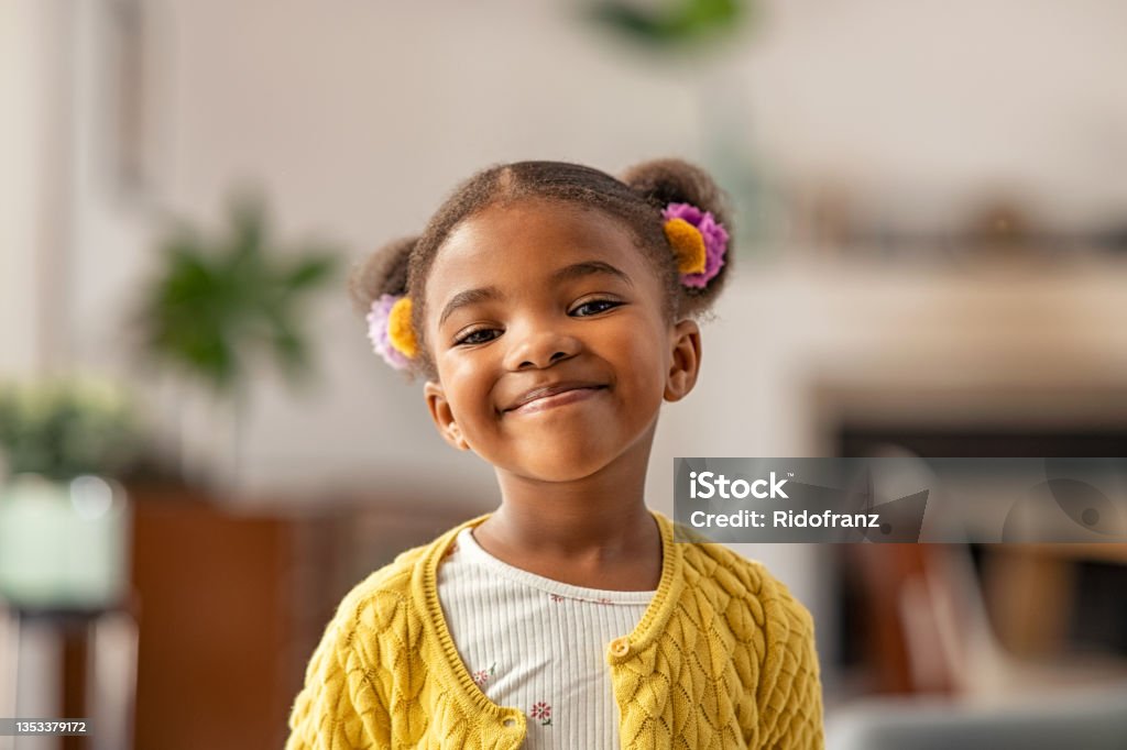 Cute little african american girl looking at camera Smiling cute little african american girl with two pony tails looking at camera. Portrait of happy female child at home. Smiling face a of black 4 year old girl looking at camera with afro puff hair. Child Stock Photo