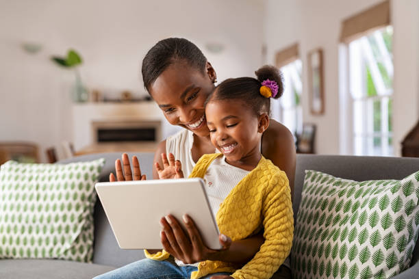 Cute little daughter and african mother using digital tablet to do a video call Smiling ethnic mother and funny daughter on video call using digital tablet at home. African american mature mother and little girl using digital tablet while making video call and waving hello sitting on sofa. Happy black woman and her cute little kid sitting on sofa and doing video conference with grandparents. black mother and adult daughter stock pictures, royalty-free photos & images