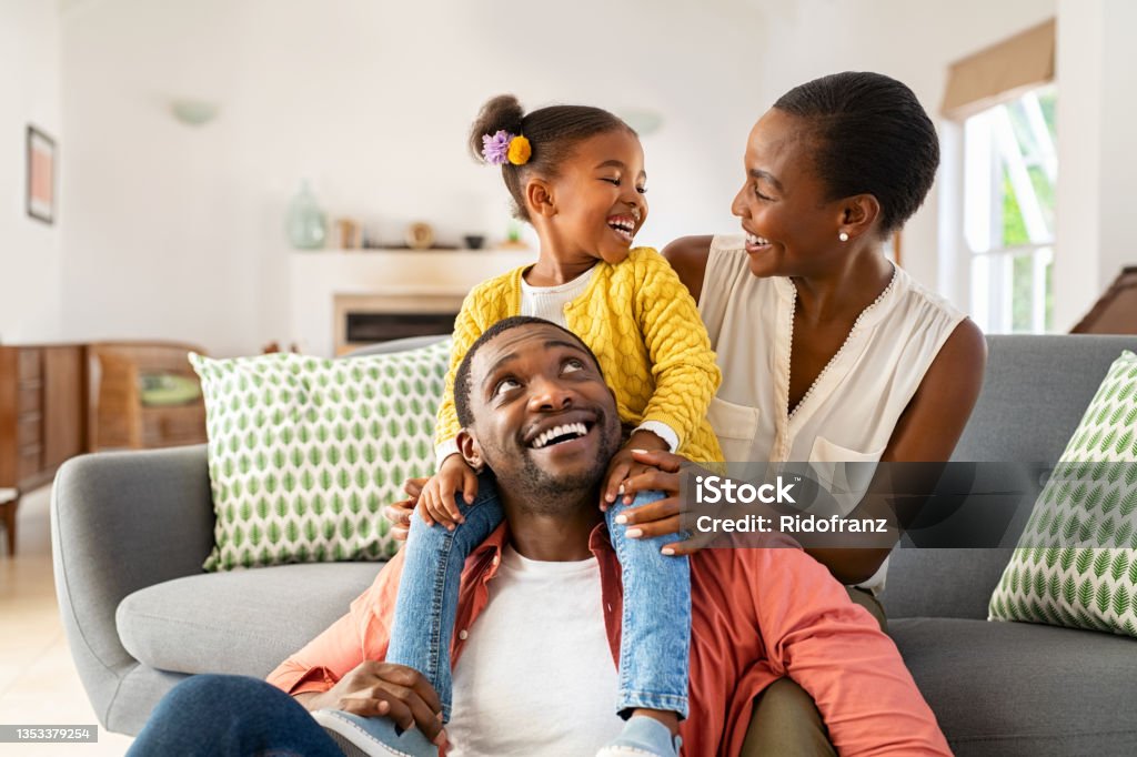 Little black girl playing with parents at home Cheerful little girl sitting on father shoulder while playing with mother at home. Happy black  family enjoying weekend at home. Cute little daughter sitting on fathers shoulder and play with her mom. Family Stock Photo