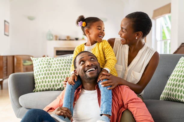Little black girl playing with parents at home Cheerful little girl sitting on father shoulder while playing with mother at home. Happy black family enjoying weekend at home. Cute little daughter sitting on fathers shoulder and play with her mom. african american stock pictures, royalty-free photos & images
