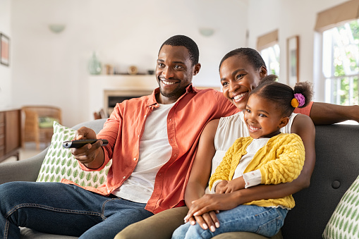 Relaxed african american family watching TV together. Happy mature father changing television channel using remote control with daughter sitting on mother's lap at home. Cheerful black family watching movie together and having fun.