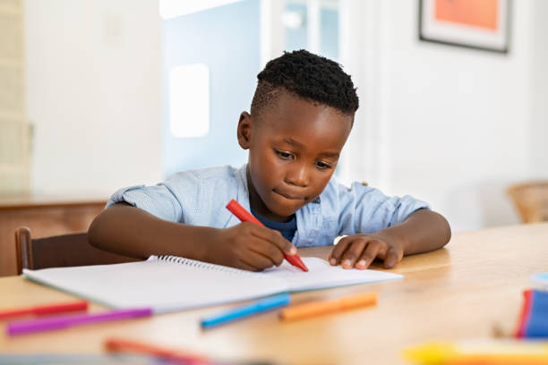 Little boy paints in notebook at home Little black boy sitting on table and painting with colored marker on book. Cute african american child drawing in the living room at home. Schoolboy colouring in book with red marker at home. young black boys stock pictures, royalty-free photos & images