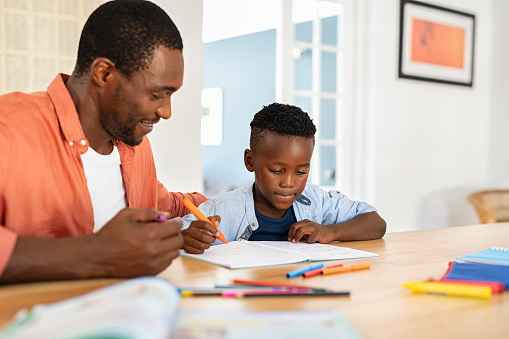 Mid black father helping his son with homework. African american boy studying with dad at home. Cute little child doing homework and studying with the help of his mature dad.