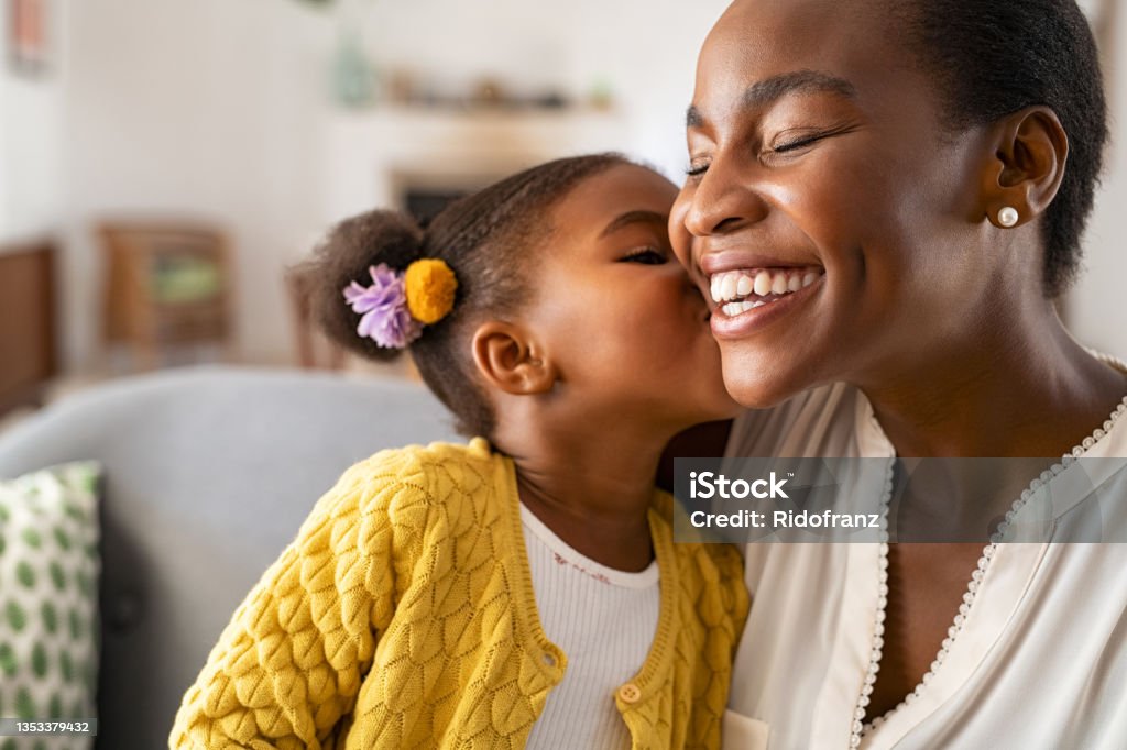 Cute little girl kissing mother on cheek Close up of beautiful daughter kissing mother on cheek at home. African american little girl giving kiss to happy mother. Lovely black female child kissing cheerful and proud woman on cheek with copy space for mother's day. Mother's Day Stock Photo