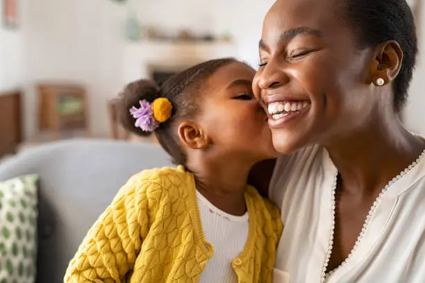 Close up of beautiful daughter kissing mother on cheek at home. African american little girl giving kiss to happy mother. Lovely black female child kissing cheerful and proud woman on cheek with copy space for mother's day.