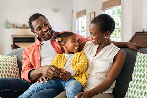 Smiling mature black family relaxing on couch. Happy middle aged man and beautiful woman sitting on sofa at home with daughter while talking together. Happy african american father and mother with one little cute girl relaxing on couch cuddling.