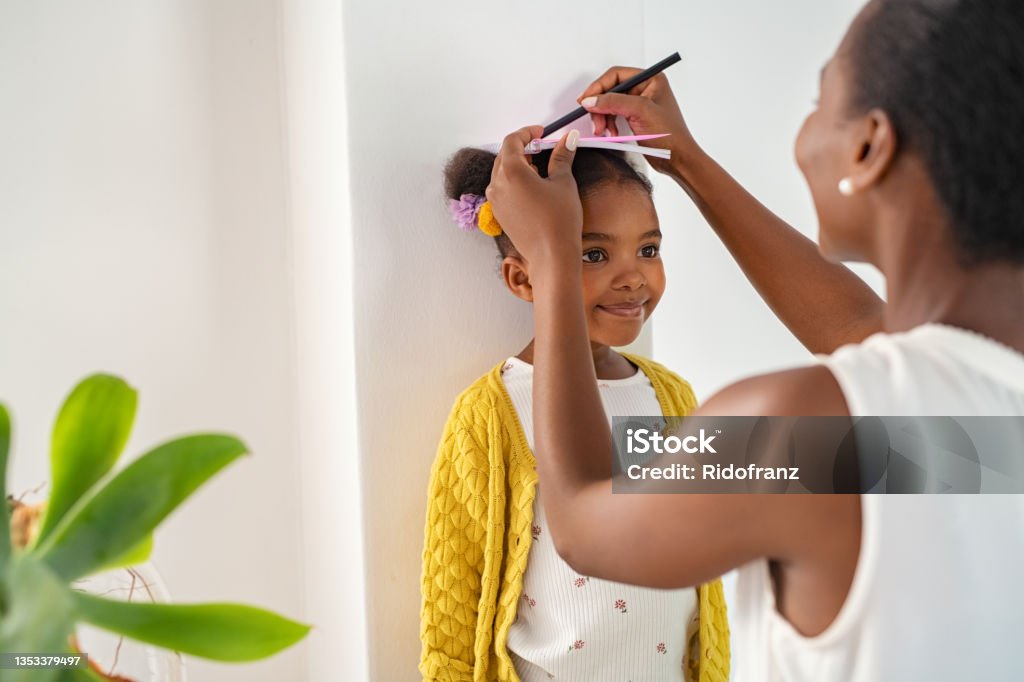 Mother measuring daughter's height and marking on wall at home African middle aged mother measuring daughter height and marking it on wall at home. Happy black mother measuring cute little girl height at home using book and pen. Cheerful mature woman measuring her daughter's progression against white wall. Child Stock Photo