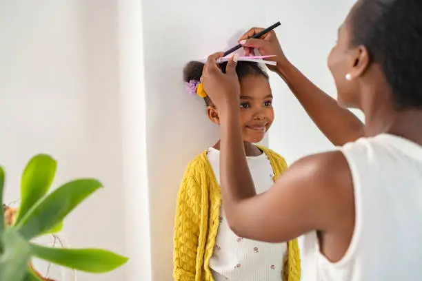 African middle aged mother measuring daughter height and marking it on wall at home. Happy black mother measuring cute little girl height at home using book and pen. Cheerful mature woman measuring her daughter's progression against white wall.