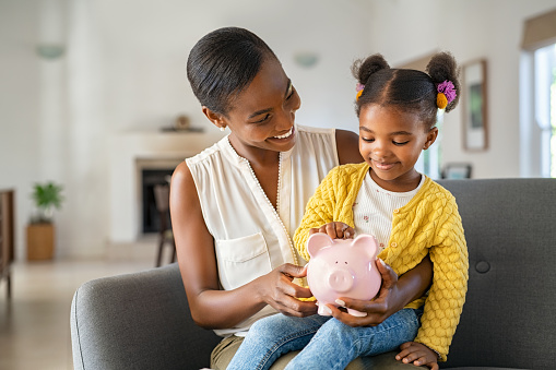 Smiling mature african american mother helping daughter sitting on lap putting money in piggy bank. Cute little black girl saving money by adding a coin in piggy bank with mother at home. Happy daughter sitting on mom