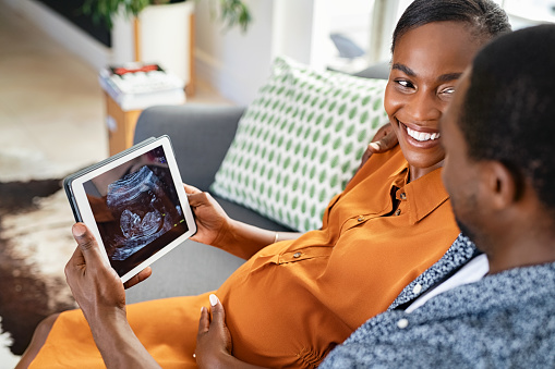 Black mature pregnant couple watching ultrasound picture on digital tablet. Smiling black pregnant woman looking at husband while feeling happy and excited on seeing baby ultrasound on digital tablet. Pregnant mature woman looking her baby on the tablet with her husband while sitting on couch at home.