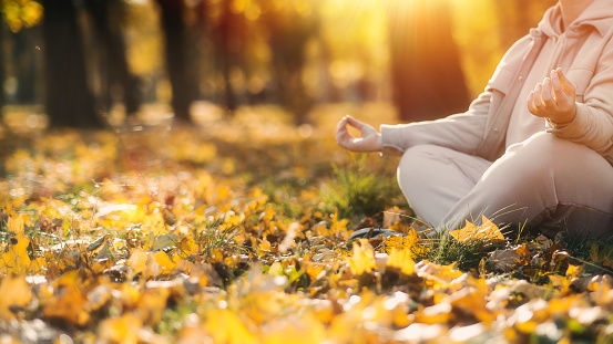 Caucasian middle aged woman meditating in lotus pose at autumn park with sunlight. Yoga at fall park. Woman in glasses relaxing on yellow leaves. Meditation, Mental health, self care, mindfulness.